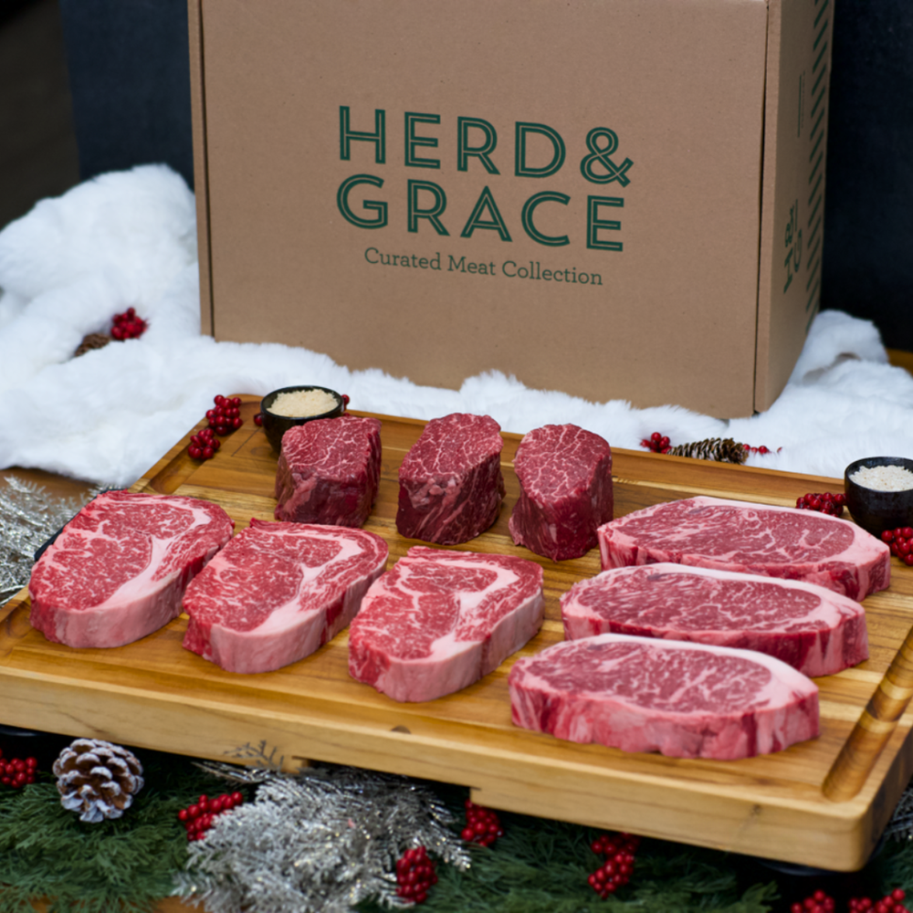 Raw marbled beef steaks arranged on a wooden board in front of a large cardboard box labeled HERD & GRACE Curated Meat Collection, styled with white faux fur, pinecones, berries, and holiday greenery.