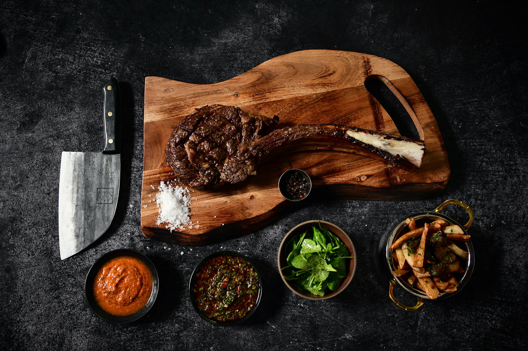 Wood cutting board with sliced steak, chef knife, salt, peppercorn bowl, and sauce bowls on dark surface