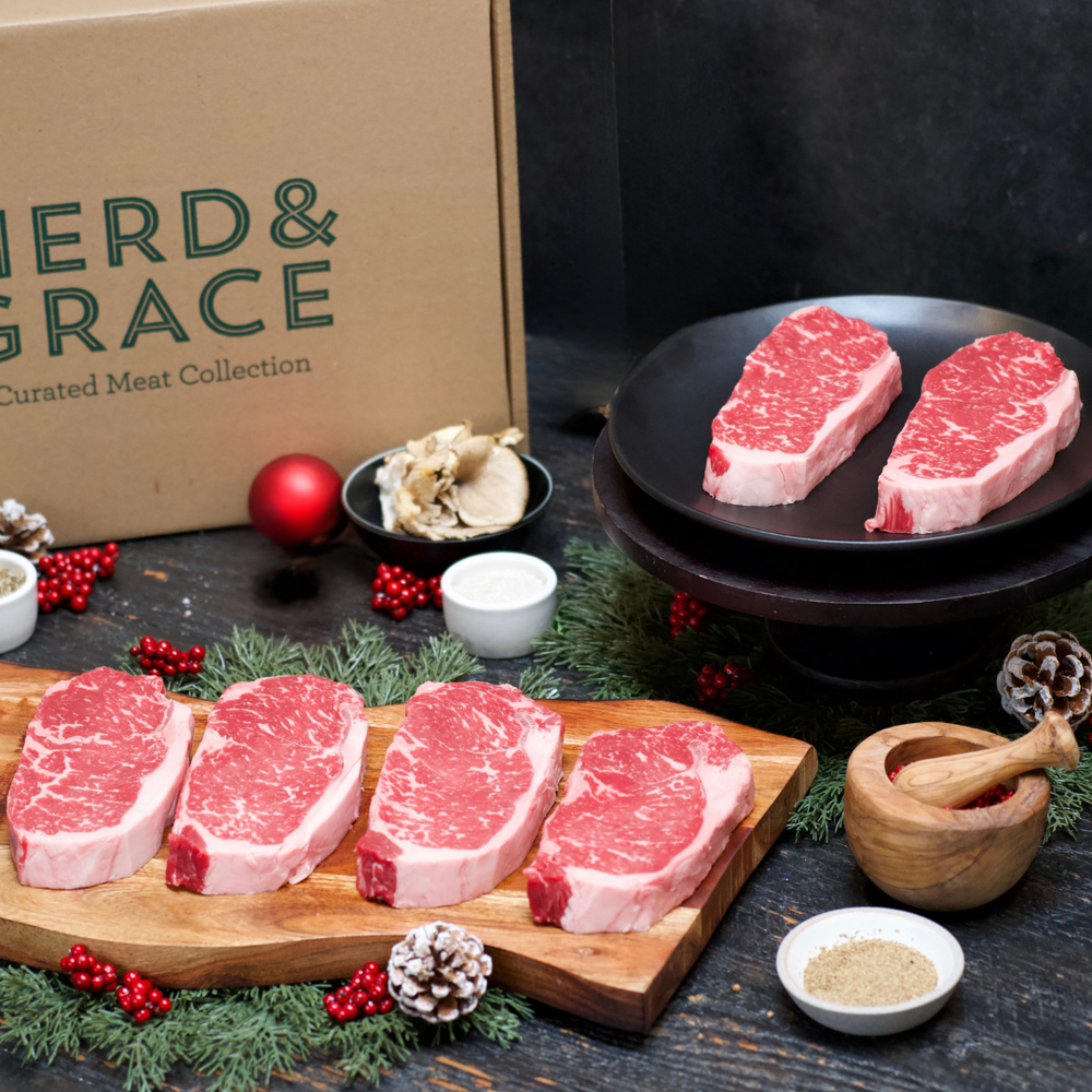 Four raw marbled beef steaks arranged on a wooden board with holiday greenery and red berries, alongside two more steaks on a black plate. A kraft box labeled Herd & Grace is visible in the background, along with small bowls of seasoning and ingredients on a dark tabletop.