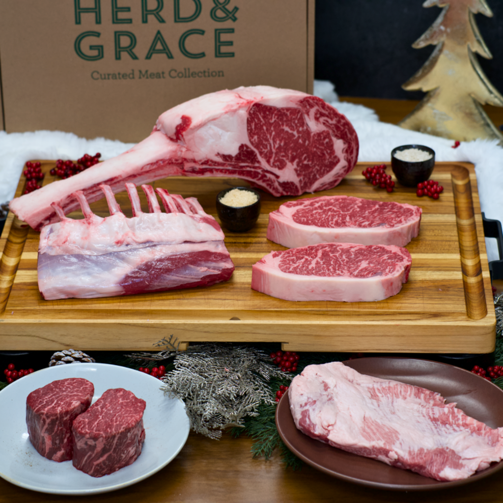 A curated meat collection display featuring multiple raw beef cuts arranged on a wooden cutting board and plates, with a branded Herd & Grace box in the background and holiday styling with red berries and evergreen branches.