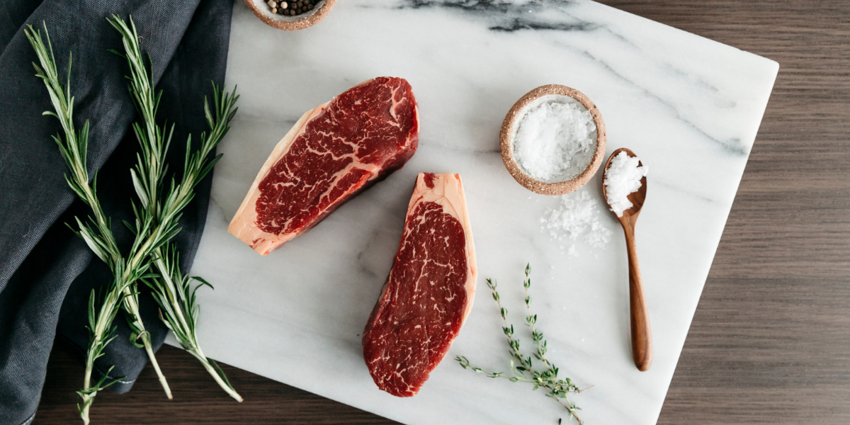 Two raw steaks on a marble surface with rosemary, salt, and pepper.